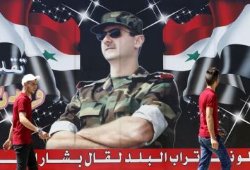 Syrian youths walk past a billboard showing a picture of Syrian President Bashar al-Assad wearing sunglasses while dressed in a Field Marshal's camouflage fatigues, on display in the centre of the capital Damascus on July 9, 2018, with a caption below reading in Arabic: "If the country's dust speaks, it will say Bashar al-Assad." / AFP PHOTO / LOUAI BESHARA