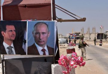 Members of Russian and Syrian forces stand guard near posters of Syrian President Bashar al-Assad and his Russian counterpart Vladimir Putin at the Abu Duhur crossing on the eastern edge of Idlib province on August 20, 2018. - Civilians are coming from rebel-held areas in Idlib province and entering regime-held territories through the Abu Duhur crossing, some of them returning to their villages that were recaptured by the regime forces earlier this year. (Photo by George OURFALIAN / AFP)